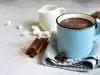 Imagem de uma caneca azul com chocolate quente. Na foto, pedaços de canela em pau, anis estrelado e uma mini jarra branca.